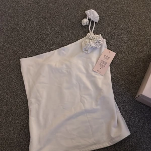 Vit one shoulder topp från Nelly - Supersnygg vit kort topp från Nelly med one shoulder och axelband dekorerat med coola tygrosor. Klänningen har en clean och stilren look, perfekt för dig som gillar trendiga detaljer och enkel design.