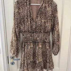 Zara klänning - Snygg kortklänning med leopardmönster och v-ringning. Klänningen har långa ärmar med resår vid handlederna och markerad midja med smock. Volangdetalj nertill ger extra flöde. Perfekt för dig som vill sticka ut med en trendig look. Litet hål i armhålan, går att laga lätt