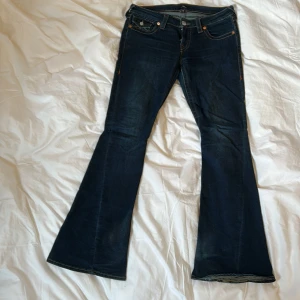 True Religion bootcut jeans mörkblå - Mörkblå bootcut jeans från True Religion med ikoniska bakfickor med kontrastsömmar och silverfärgade knappar. Klassisk femficksmodell i jeansmaterial med orange sömmar och låg midja. Perfekta för dig som gillar en retro vibe och markerade detaljer. Model Joey. Lite slita nedtill kan så priset kan därför diskuteras, nypris 1200kr mitt pris 800kr.