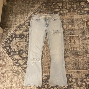 Ljusblå lågmidjade bootcut jeans från Zara - Säljer ett par ljusblå jeans från Zara i bootcut-modell med slitna detaljer på låret och knät. Jeansen har klassisk femficksdesign och är tillverkade i bomull. Perfekta för en avslappnad och trendig look.