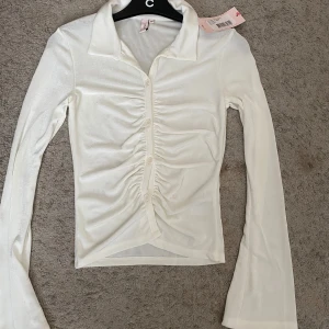 Vit glansig blus med rynk och knappar - Snygg vit blus i glansigt material med lång ärm, krage och knappar framtill. Rynkade detaljer längs knappraden ger en cool och trendig look. Perfekt att styla med jeans eller kjol för en fräsch outfit.