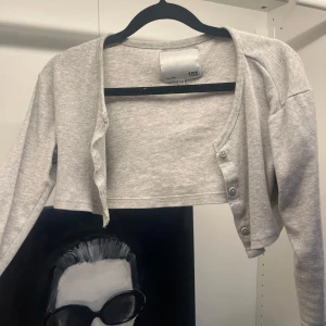 Grå croppad kofta med knappar - Grå croppad kofta (bolero)från 157 med ribbad struktur och långa ärmar. Koftan har knappar framtill och en enkel, stilren design som passar perfekt till lager-på-lager. Superenkel att matcha med både jeans och kjol.