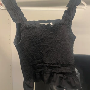 Svart smockad linnetopp med volang - Supersöt svart linnetopp med breda smockade axelband och smockad överdel. Toppen har en volangkant längst ner som ger en extra touch. Perfekt att styla med jeans eller kjol för en trendig look.