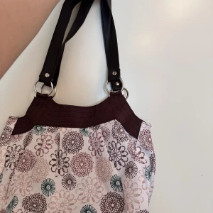 Blommig axelväska med bruna detaljer Y2K - !Buda!Söt axelväska med blommigt mönster i brunt, beige och grönt på ljus botten. Väskan har breda mörkbruna axelband i syntet och silverfärgade ringdetaljer. Stängs med magnetknapp och har ett rymligt foder i brunt tyg. Perfekt för dig som gillar unika accessoarer.