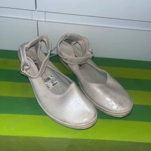 Vita ballerinaskor med rem från Dinsko - Snygga vita ballerinaskor från Dinsko med diskret ormskinnsmönster och justerbar rem runt vristen. Skorna har rund tå och en liten platåsula som ger extra komfort. Perfekta för dig som gillar stilrena och feminina skor.