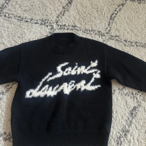 Svart Saint Laurent stickad tröja - Svart stickad tröja från Saint Laurent med vit, fluffig logotyp framtill. Rund halsringning och ribbade muddar vid ärmslut och nederkant. Perfekt för dig som gillar statement-plagg och exklusiv streetstyle. Storlek S, men smalare M passar också. Skambud ingnoreras och eventuellt blockeras. Fraktar samma dag vid köp innan 20:00. 