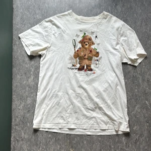 Vit Polo Bear t-shirt från Ralph Lauren - Vit t-shirt från Ralph Lauren med Polo Bear-tryck där björnen är klädd som äventyrare. Klassisk passform, rund hals och korta ärmar. Perfekt för dig som gillar coola och lekfulla prints.