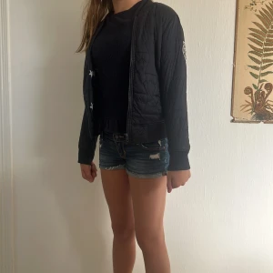 Svart quiltad bomberjacka - Svart quiltad bomberjacka med dragkedja framtill och ribbade muddar vid ärmslut och midja. Jackan har en patch på ena ärmen och klassisk bomberkrage. Perfekt för dig som vill ha en stilren och enkel jacka med snygga detaljer.