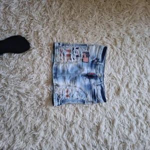 Ljusblå jeanskjol med USA-detaljer - Cool ljusblå jeanskjol med slitna detaljer, nitar och broderade amerikanska flaggor. Kjolen har klassiska fickor både fram och bak samt en knapp och dragkedja framtill. Perfekt för dig som gillar streetstyle och unika plagg.