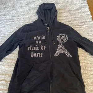 Svart hoodie med strass och tryck - Svart hoodie från BLINGLUCID i storlek XS med dragkedja, huva och snörning. Framsidan har strasstenar som bildar Eiffeltornet och texten 'paris au clair de lune'. Perfekt för dig som gillar statement-plagg med Paris-vibe.