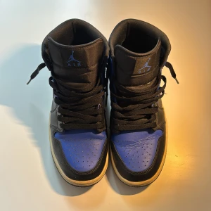 Nike Air Jordan 1 Mid OG Svart/Blå Royals - Säljer ett par Nike Air Jordan 1Mid OG i svart och blått skinn med klassisk vita detaljer och svarta snören. Ikonisk siluett med högt skaft, perforerad tå och tydlig Swoosh på sidan. Perfekta för dig som vill sticka ut med en tidlös sneaker.