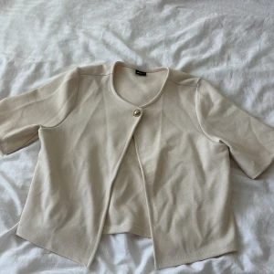 Beige cardigan jacka från Gina Tricot - Kort beige cardigan jacka från Gina Tricot med rund halsringning och en snygg guldig knapp framtill. Jackan har korta ärmar och är gjord i ett mjukt, stickat material. Perfekt för att lyfta en enkel outfit med en clean och stilren look.