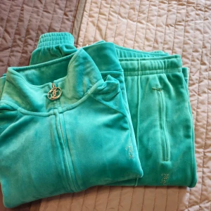 Juicy Couture grönt set  - Grönt set från Juicy Couture med glittrande stenar på ryggen och rumpan där det står 'Juicy'. Tröjan har dragkedja med JC-logga, långa ärmar och ribbade muddar samt ficka med dragkedja. Perfekt för dig som vill ha den ikoniska Y2K-looken. Byxorna har sydda pressveck och fickor med dragkedja