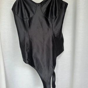 Svart satin korsettbody - Snygg svart korsettinspirerad body i glansigt satinliknande material. Toppen har markerade kupor och smala axelband, samt knäppning i grenen. Perfekt för dig som vill ha en edgy och trendig look.