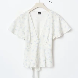 Vit blommig omlottopp med knytband - Supersöt vit omlottopp från Gina Tricot med ljusblått blommönster. Toppen har v-ringning, korta ärmar och knytband i midjan som ger en fin siluett. Perfekt för vår och sommar när du vill ha en fräsch och romantisk look. (Den är knappt använd) 