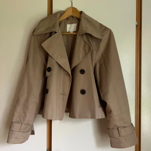 Kort beige trenchcoat från HM - Snygg croppad trenchcoat i beige med bred krage, dubbelknäppning och bältdetaljer vid ärmsluten. Sparsamt använd i strl S (normal i storleken)