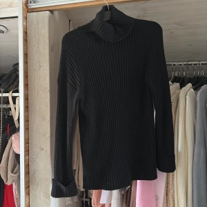 Svart ribbad polotröja - Snygg svart polotröja med ribbat mönster och hög krage. Tröjan är långärmad och har en klassisk, stilren look som passar perfekt till jeans eller kjol. Materialet känns mjukt och stretchigt, vilket gör den bekväm att bära hela dagen.