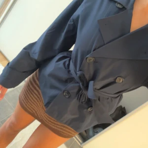 Mörkblå trenchcoat med bälte - Snygg mörkblå trenchcoat med dubbelknäppning och bälte i midjan. Jackan har klassisk krage, långa ärmar och stora knappar framtill. Perfekt för dig som vill ha en stilren och tidlös look.