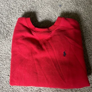 Röd tröja från Polo Ralph Lauren - Klassisk röd tröja från Polo Ralph Lauren med rund halsringning och broderad marinblå logga på bröstet. Tröjan har en enkel och stilren design, perfekt för dig som gillar en clean look. Tillverkad i mjuk bomull för skön känsla.