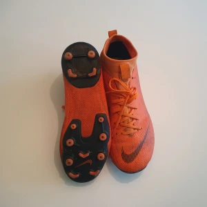 Orange Nike fotbollsskor med snörning - Snygga orangea fotbollsskor från Nike med svart swoosh-logga på sidan. Skorna har snörning, plös med flik och högre skaft för extra stöd. Ovandelen är i syntetmaterial med mönstrad yta och sulan har fasta dobbar för gräsplan.