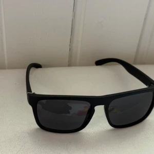 Svarta polariserade solglasögon - Snygga svarta solglasögon med rektangulära bågar och polariserade mörka glas. Klassisk och stilren design som passar till det mesta. Bågarna är i matt svart plast och har en liten metall-detalj vid gångjärnet där det står 'Polarized'.