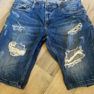 Blå slitna jeansshorts från FSBN - Riktigt snygga blå jeansshorts från FSBN med slitna detaljer och stora hål framtill för en cool, avslappnad vibe. Klassisk femficksmodell, normal passform och råa kanter. Perfekta för sommardagar när du vill sticka ut lite extra.