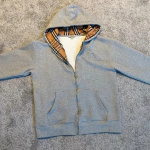 Grå hoodie från Burberry med dragkedja - Snygg grå hoodie från Burberry med klassiskt rutigt foder i huvan. Tröjan har dragkedja framtill, två fickor och en skön huva. Perfekt för dig som gillar stilrena och bekväma plagg med en touch av lyx.