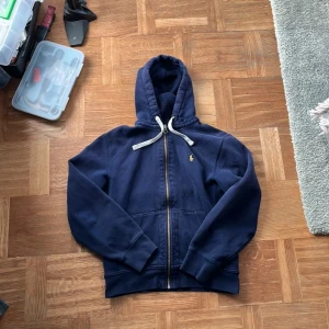 Mörkblå huvtröja från Ralph Lauren - Säljer en snygg mörkblå huvtröja från Ralph Lauren. Den har en dragkedja framtill och en liten broderad logga på bröstet. Tröjan är i bra skick och perfekt för höst och vår. Den har långa ärmar och en skön huva med dragsko.