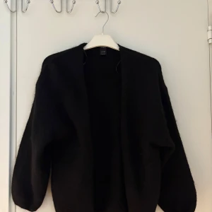 Svart stickad kofta från Lindex - Mysig svart stickad kofta från Lindex Koftan har lång ärm, öppen framtill och är fluffig i materialet. Perfekt att slänga på sig när det är kyligt ute. Enkel och stilren design som funkar till många outfits.