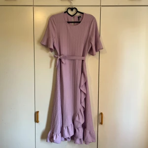 Ljuslila volangklänning med knytband - Superfin ljuslila långklänning från Vero Moda med volangdetaljer längs fållen och ärmarna. Klänningen har ett knytband i midjan och lätt struktur i tyget. Skriv för fler bilder eller funderingar💜