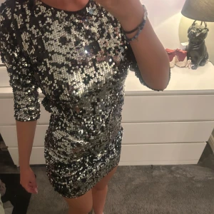Glittrig silver paljettklänning - Säljer en kort, tight klänning täckt av silvriga paljetter. Klänningen har trekvartsärm, öppen rygg och dragkedja bak. Perfekt för dig som vill sticka ut och glänsa på festen! Sitter figurnära och har en riktigt snygg cut out-detalj i ryggen.