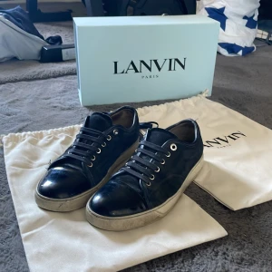 Lanvin Captoe - Snygga svarta sneakers från Lanvin i glansigt skinn med rund tå och vita sulor. Klassisk modell med snörning och diskreta detaljer. Perfekta för dig som gillar stilrena och exklusiva sneakers med lyxig känsla. Slitna undertill 