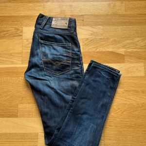 Replay jeans i mörkblå bomull - Snygga Replay jeans i mörkblå tvätt med klassiska kontrastsömmar och detaljer i rött. Jeansen är tillverkade i 100% bomull och har raka ben samt fem fickor. Perfekta för dig som gillar en tidlös och stilren look. 30/32