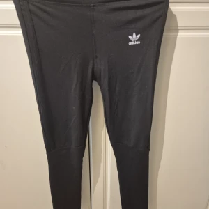 Svarta leggings från Adidas strl M - Svarta leggings från Adidas med klassisk logga på höften. Tight passform och stretchigt material som sitter skönt. Perfekta för träning eller chill, med diskreta sömmar och hög midja för en snygg siluett.