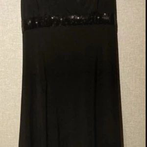 Svart långklänning med paljettdetalj - Elegant svart långklänning från ICHI med en glittrig paljettrad vid midjan. Klänningen har en stilren och klassisk siluett som faller snyggt längs kroppen. Perfekt för dig som vill ha en tidlös look med lite extra bling.