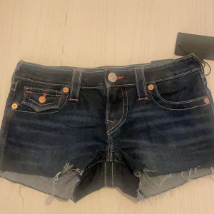 True Religion jeansshorts low rise - Snygga mörkblå jeansshorts från True Religion med låg midja och råa, fransiga benslut. Modellen heter Joey Cut-Off och har coola broderade bakfickor med logga och röda detaljer. Klassisk femficksdesign och silverfärgade knappar. Perfekta för sommaren! 💖