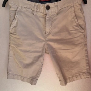Beige shorts från Superdry, 28W - Snygga beige shorts från Superdry International med klassisk design och raka ben. De har fickor både fram och bak, bälteshällor och knappstängning. Perfekta för chill dagar och sommarhäng, tillverkade i mjuk bomull som känns skön mot huden.