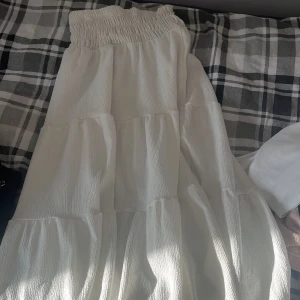 Vit volangkjol med resårmidja - Superfin vit långkjol med flera volanger och smockad resår i midjan. Kjolen har en luftig och flowy känsla, perfekt för varma dagar. Materialet är lätt och har en snygg struktur som ger extra liv till outfiten.