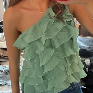 Grön volangig oneshoulder topp - Säljer en mintgrön topp med flera lager volanger och oneshoulder-design. Toppen är ärmlös på ena sidan och har en luftig, flowy känsla som gör den extra snygg till jeans eller kjol. Aldrig använd! 