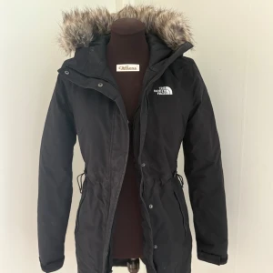 Svart parkajacka från The North Face - Svart parkajacka från The North Face med fuskpäls på huvan och dragsko i midjan. Jackan har långa ärmar, dragkedja och knappar framtill samt två fickor med dragkedja. Perfekt för kalla dagar när du vill hålla stilen.