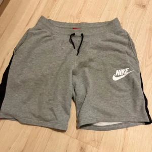 Grå Nike shorts med snörning M - Snygga grå shorts från Nike med svart snörning i midjan och vit Nike-logga på framsidan. De har svarta mesh-detaljer på sidorna och en bakficka i mesh. Perfekta för träning eller chill. Mjukt material som känns skönt mot huden.