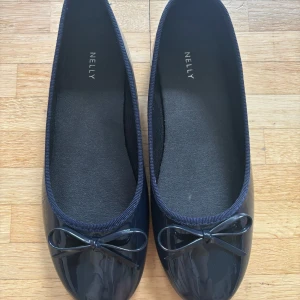 Mörkblå ballerinaskor med rosett från Nelly - Snygga mörkblå ballerinaskor från Nelly med glansig finish och söt rosettdetalj framtill. Skorna har rund tå och platt sula, perfekta för en stilren look. Tillverkade i syntetmaterial och passar till många outfits.