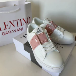 Valentino Rockstud Open sneakers vit/rosa - Snygga Valentino Rockstud Open sneakers i vitt skinn med bred rosa rem över vristen dekorerad med ikoniska nitar. Klassisk rund tå, platt sula och vita snören. Perfekta för dig som vill ha en lyxig och trendig look med coola detaljer.
