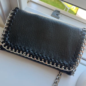 Svart axelväska med kedjedetaljer från Tiamo - Snygg svart axelväska från Tiamo i skinnimitation med grov silverfärgad kedja runt kanterna och som axelrem. Väskan har magnetlås och ett fack med dragkedja inuti. Perfekt storlek för det viktigaste och ger en cool vibe till din outfit.