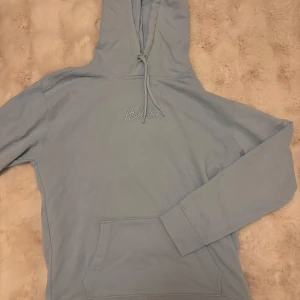 Ljusblå hoodie från Hollister - Säljer en ljusblå hoodie från Hollister med broderad logga på bröstet. Tröjan har huva med snörning, stor magficka och långa ärmar med ribbade muddar. Perfekt för chill dagar och enkel att matcha med jeans eller joggers.