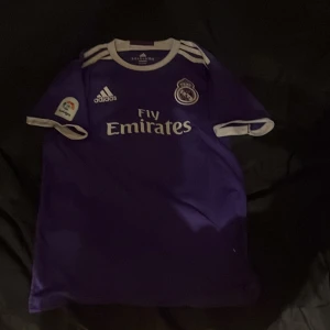 Real Madrid Ronaldo 7 lila matchtröja - Snygg lila Real Madrid fotbollströja från Adidas med Ronaldo 7 på ryggen. Tröjan har korta ärmar, vita detaljer vid krage och ärmslut, samt tryck med Fly Emirates och klubbmärke på bröstet. Materialet är lätt och andas, perfekt för träning eller match.