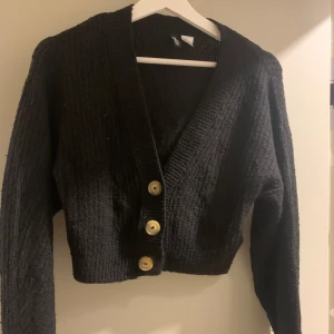 Svart stickad kofta från H&M Divided - Svart stickad kofta från H&M Divided i croppad modell med stora beige knappar framtill. Koftan har lång ärm, djup v-ringning och grovstickad struktur. Perfekt för lager-på-lager och chill vibes.