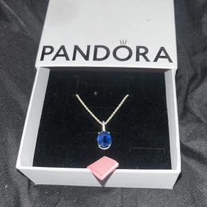 Pandora halsband med blå sten - Snyggt halsband från Pandora med en silverkedja och en oval, intensivt blå sten som hänger i mitten. Stenen är infattad i en glittrande ram som ger extra lyxig känsla. Perfekt för dig som gillar stilrena smycken med färgklick.