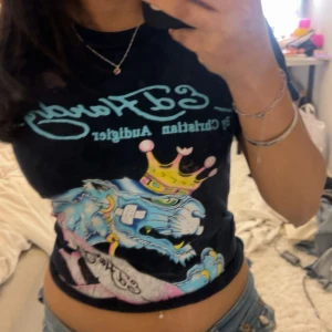  Ed Hardy t-shirt med tryck - Säljer en svart t-shirt från Ed Hardy med stort färgstarkt tryck på framsidan. Motivet är en blå tiger med krona och rosa detaljer, samt Ed Hardy-logga i turkos. Modellen är kortärmad och har en normal passform. Materialet är mjuk bomull, perfekt för en cool streetstil.pris kan diskuteras 