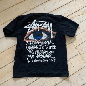 Svart Stüssy t-shirt med ögonmotiv - Svart t-shirt från Stüssy med stort grafiskt tryck på ryggen – ett blått öga och vit text i streetstil. Framtill finns en liten Stüssy-logga på bröstet. Klassisk passform och rund hals. Perfekt för dig som gillar streetwear och snygga prints.
